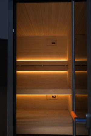 Interior of an outdoor traditional sauna with warm timber walls, soft backrest lighting and a glass door partially open.