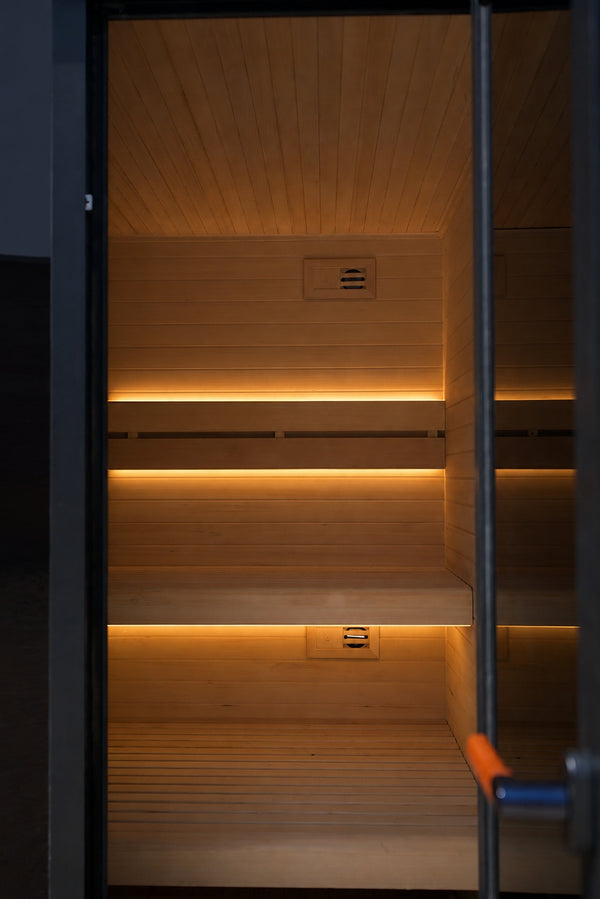 Interior of an outdoor traditional sauna with warm timber walls, soft backrest lighting and a glass door partially open.
