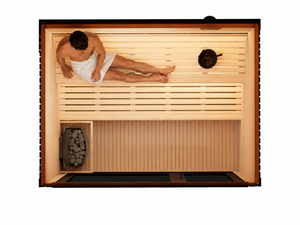 A top-down view of a wooden sauna with a man lying down inside, wrapped in a white towel. The sauna features light-colored wooden slats, a dark wood frame, a built-in firebox with gray stones, and a black hanging pot.
