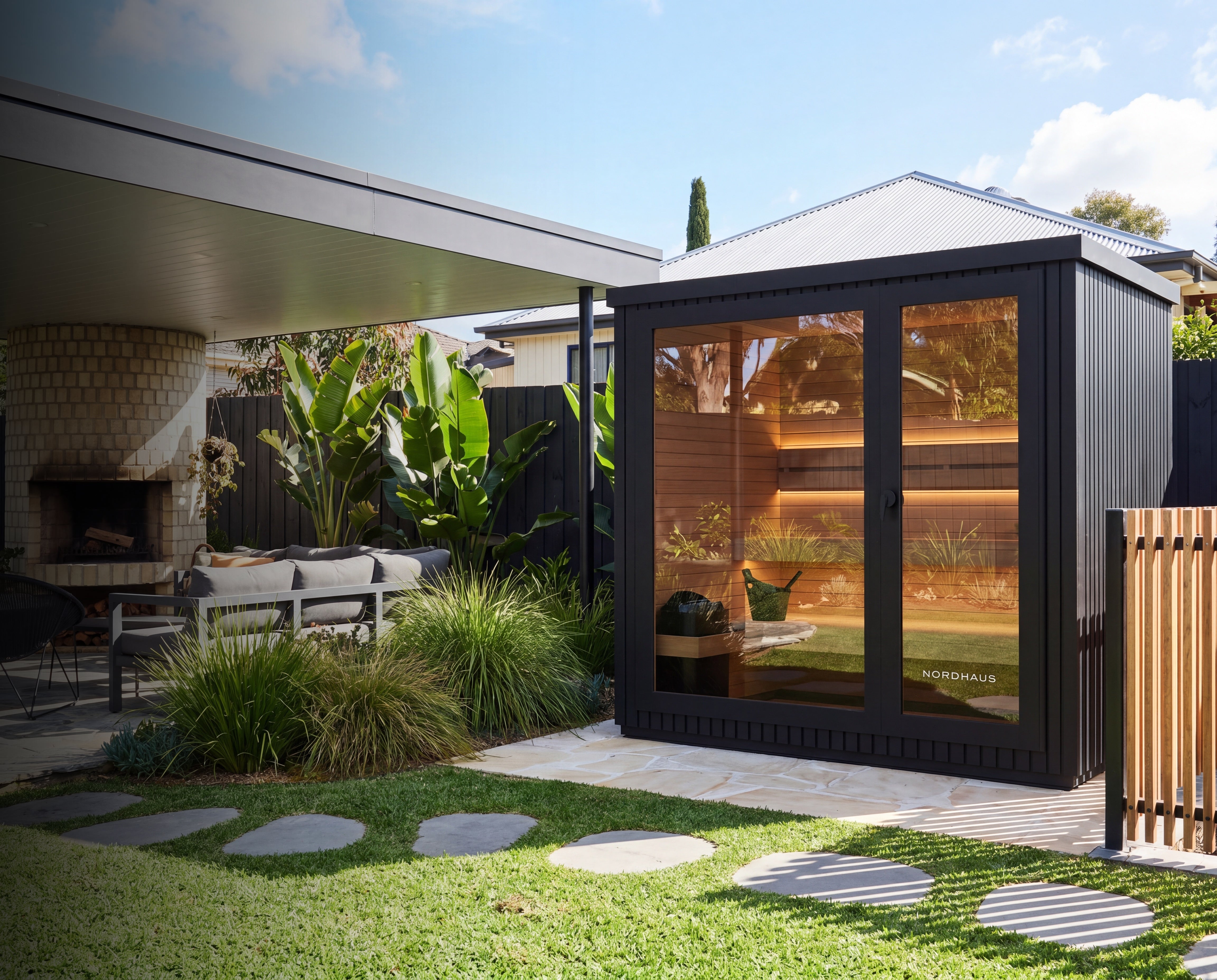 A modern black outdoor cabin with large glass doors and windows, shown in a landscaped backyard next to a patio and fireplace. The cabin interior is warmly lit, and the exterior features a dark charcoal finish with a corrugated metal roof.