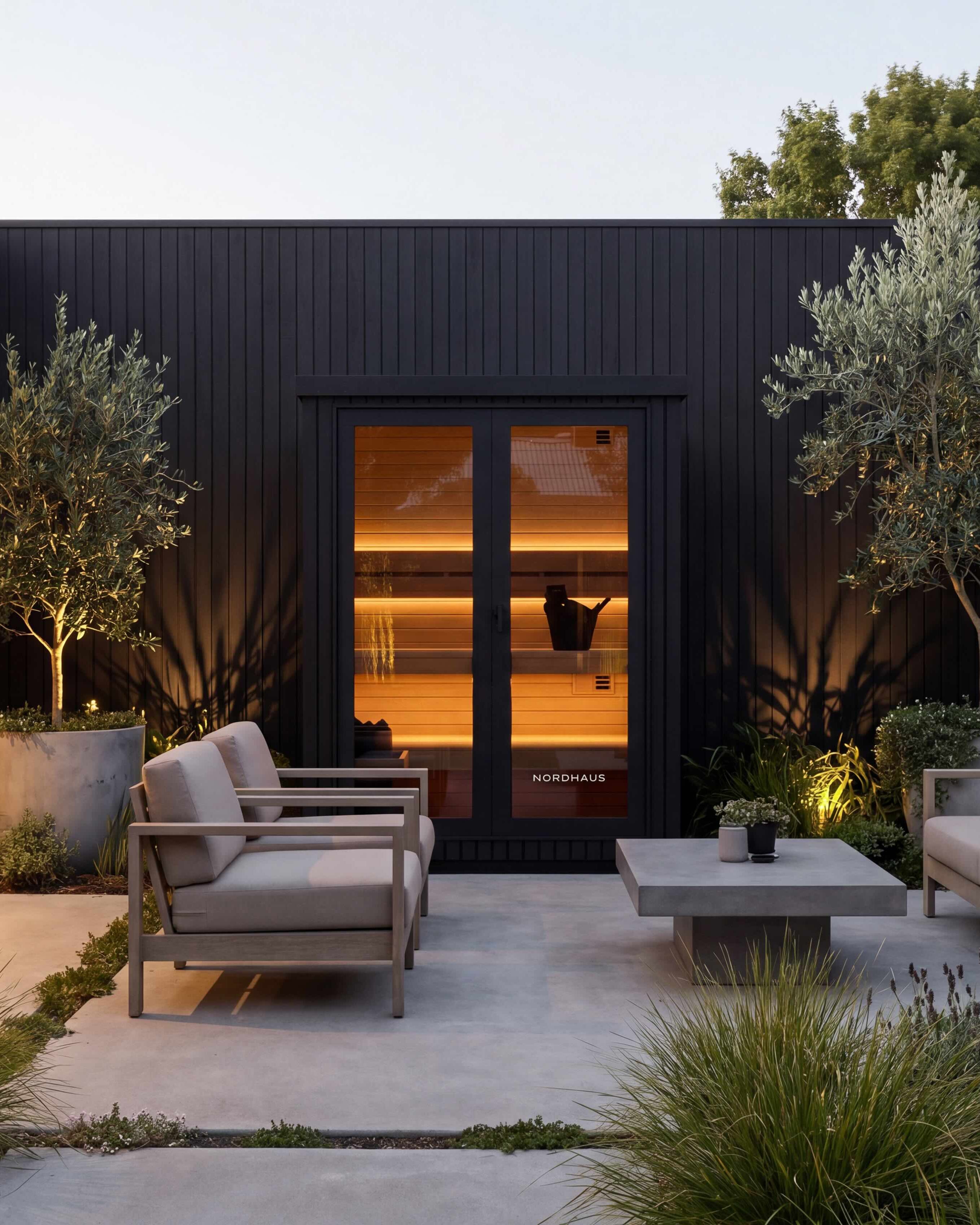 Modern outdoor patio furniture set including a sofa, two armchairs, and a low rectangular coffee table, arranged on a light stone patio in front of a black wood-clad house with olive trees and plants.