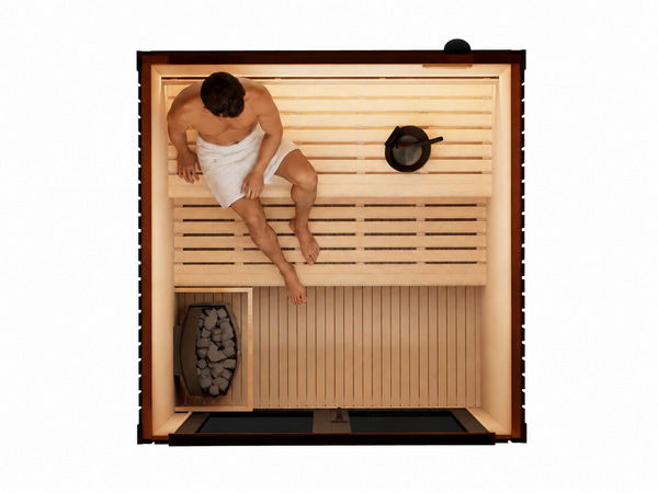Top-down view of a wooden sauna interior with a person sitting on the upper bench wrapped in a white towel, a black metal pot on the shelf, and a fire basket with logs on the lower left.