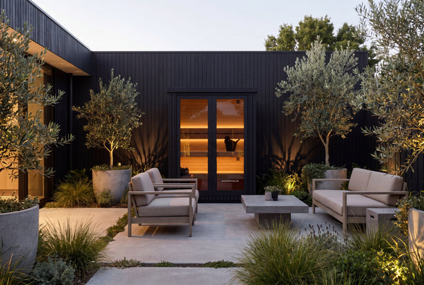Black Nordhaus outdoor traditional sauna with a warm timber interior, set within a landscaped modern courtyard with outdoor seating and olive trees.