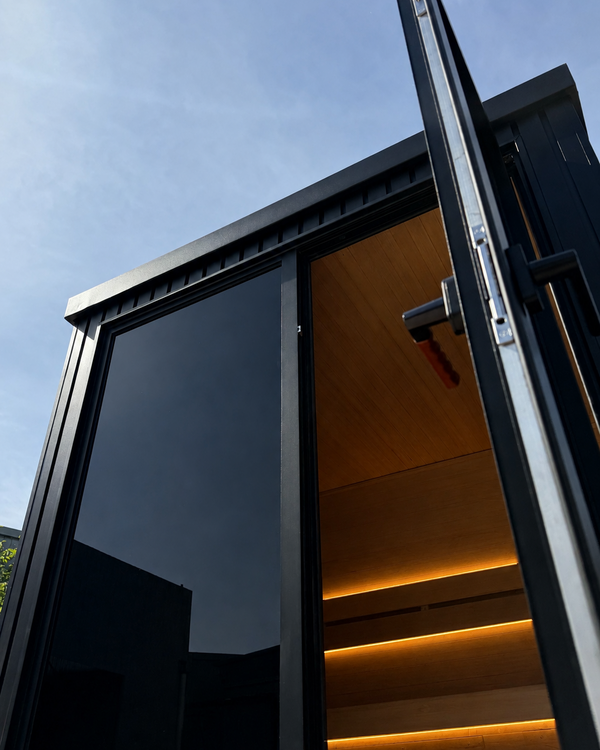 Outdoor black traditional sauna with open glass door, warm wood interior and soft backrest lighting.