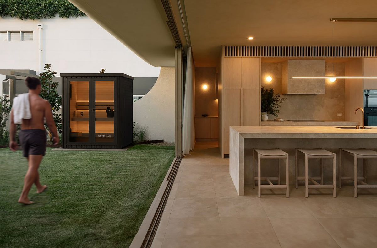 A modern outdoor indoor lifestyle scene showing a man walking on green grass next to a black glass-walled sauna cabin, with a contemporary kitchen and island in the background.